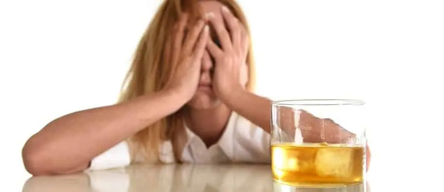 Stressed woman with face in hands beside a glass of whiskey, symbolizing alcohol addiction and emotional distress.