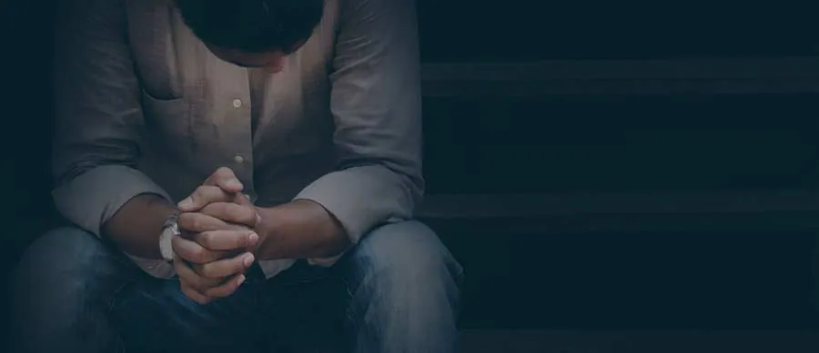 Pensive man seated alone in dim lighting, head bowed and hands clasped, symbolizing emotional struggle and fear around sobriety.