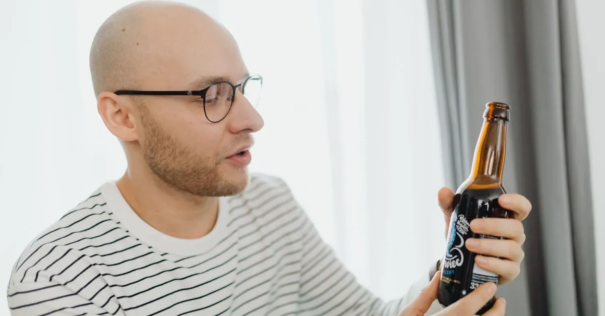 Bald man with glasses examines a bottle of craft beer indoors.