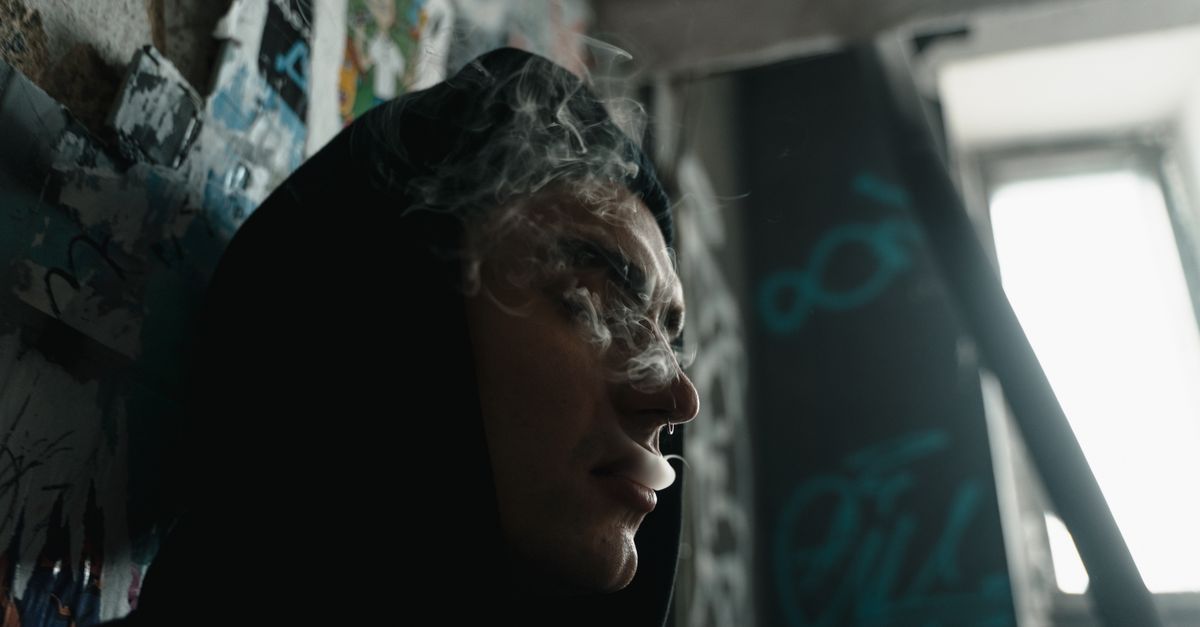 Person in hoodie surrounded by graffiti, exhaling smoke in urban setting.
