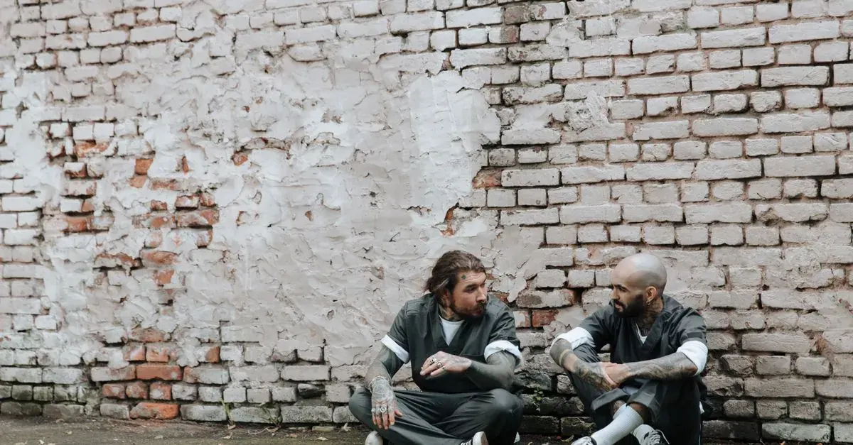 Tattooed men in prison uniforms talking while sitting outdoors against a brick wall.