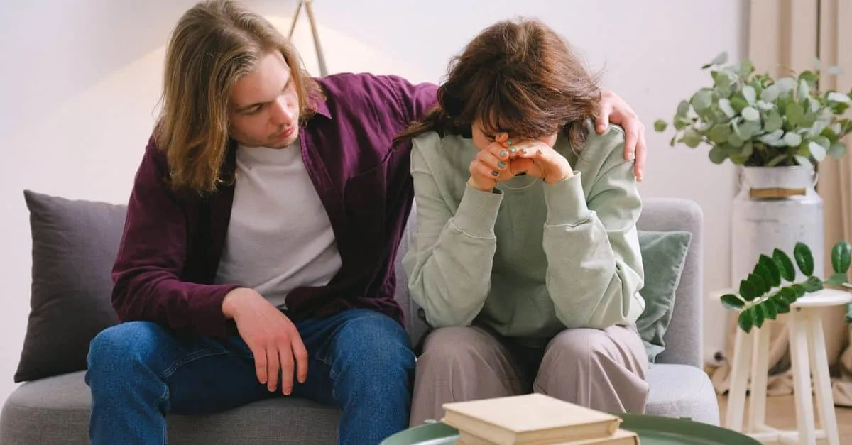 Young male in jeans comforting depressed upset female crying on gray sofa near table with books